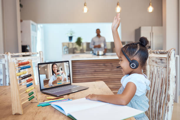 A young girl wearing a headset raises her hand during a live online class on her laptop